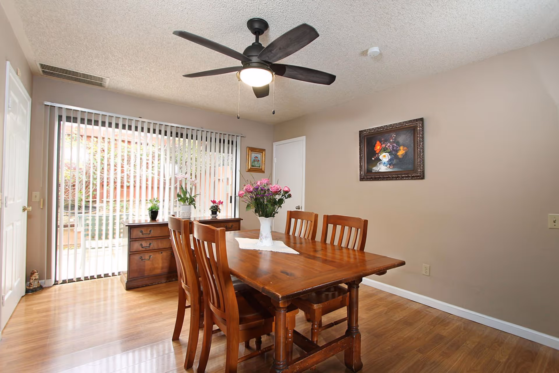 A dining room with a wooden table and six matching chairs. A vase with pink flowers is placed on a white doily in the center of the table. Behind the table is a wooden sideboard with potted plants on top. Vertical blinds cover a large window or sliding glass door, allowing natural light to enter the room. The walls are beige, with two framed paintings hanging on them. A ceiling fan with a light fixture is mounted on the ceiling.