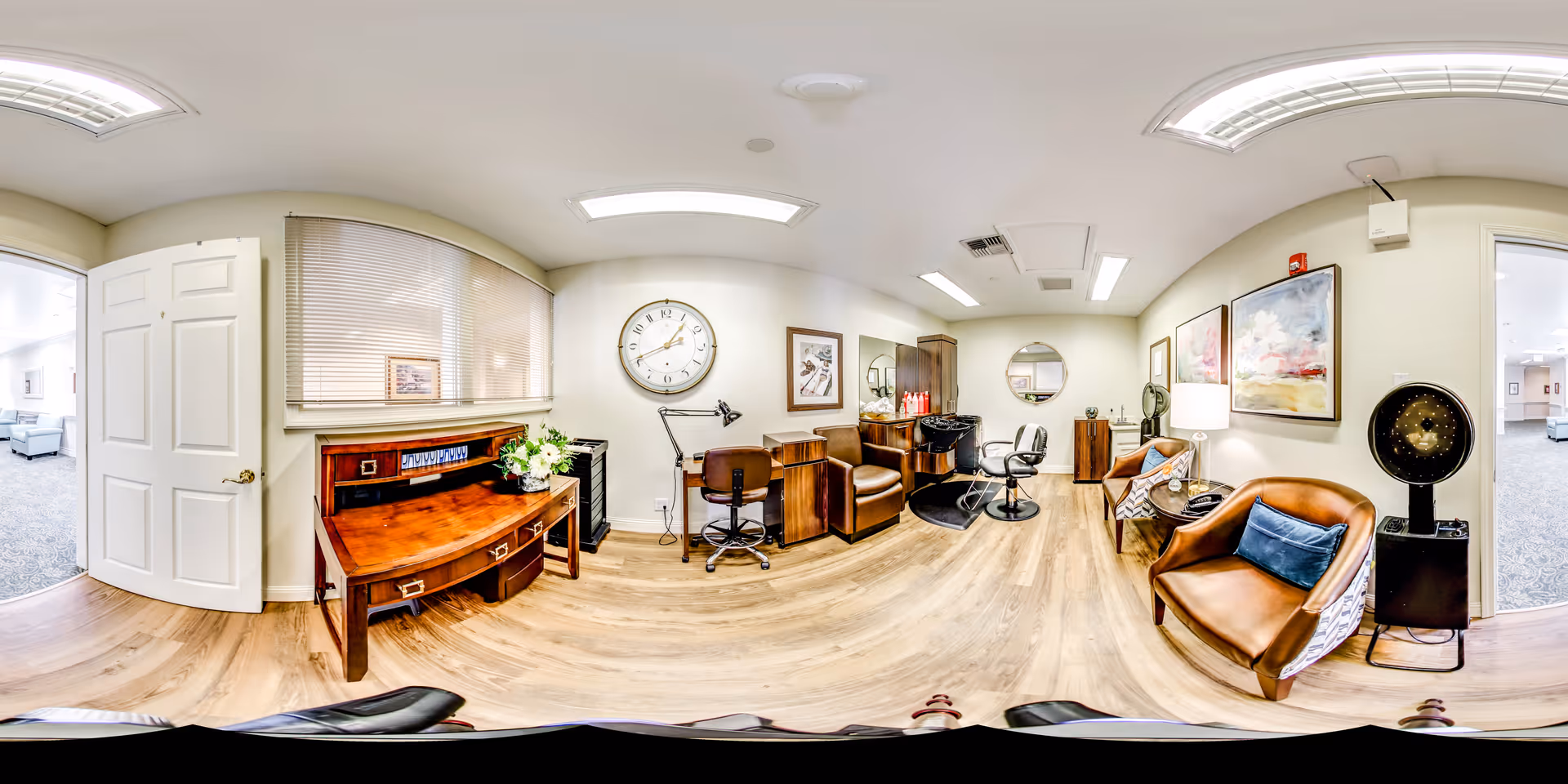 A bright, well-lit interior room with wooden flooring featuring a wooden desk with flowers, a large wall clock, two brown leather chairs, a hair washing station with a salon chair, a round mirror, and framed artwork on the walls. There are two open doorways leading to carpeted rooms with sofas visible.