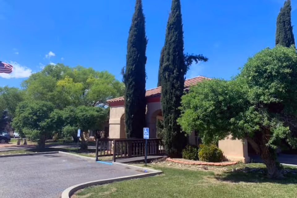 Exterior view of Desert Peaks Assisted Living and Memory Care facility showing a beige building with a red-tiled roof, surrounded by tall cypress trees and green bushes under a clear blue sky. There is a parking area with handicap parking signs and an American flag visible in the background.