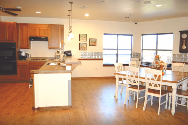 A bright kitchen and dining area with wooden floors and white walls. The kitchen features wooden cabinets, a built-in oven, a coffee maker, and a countertop with a sink and hanging pendant lights. The dining area has a wooden table with six white chairs, and windows with blinds letting in natural light. A wall clock and framed pictures are visible on the walls.