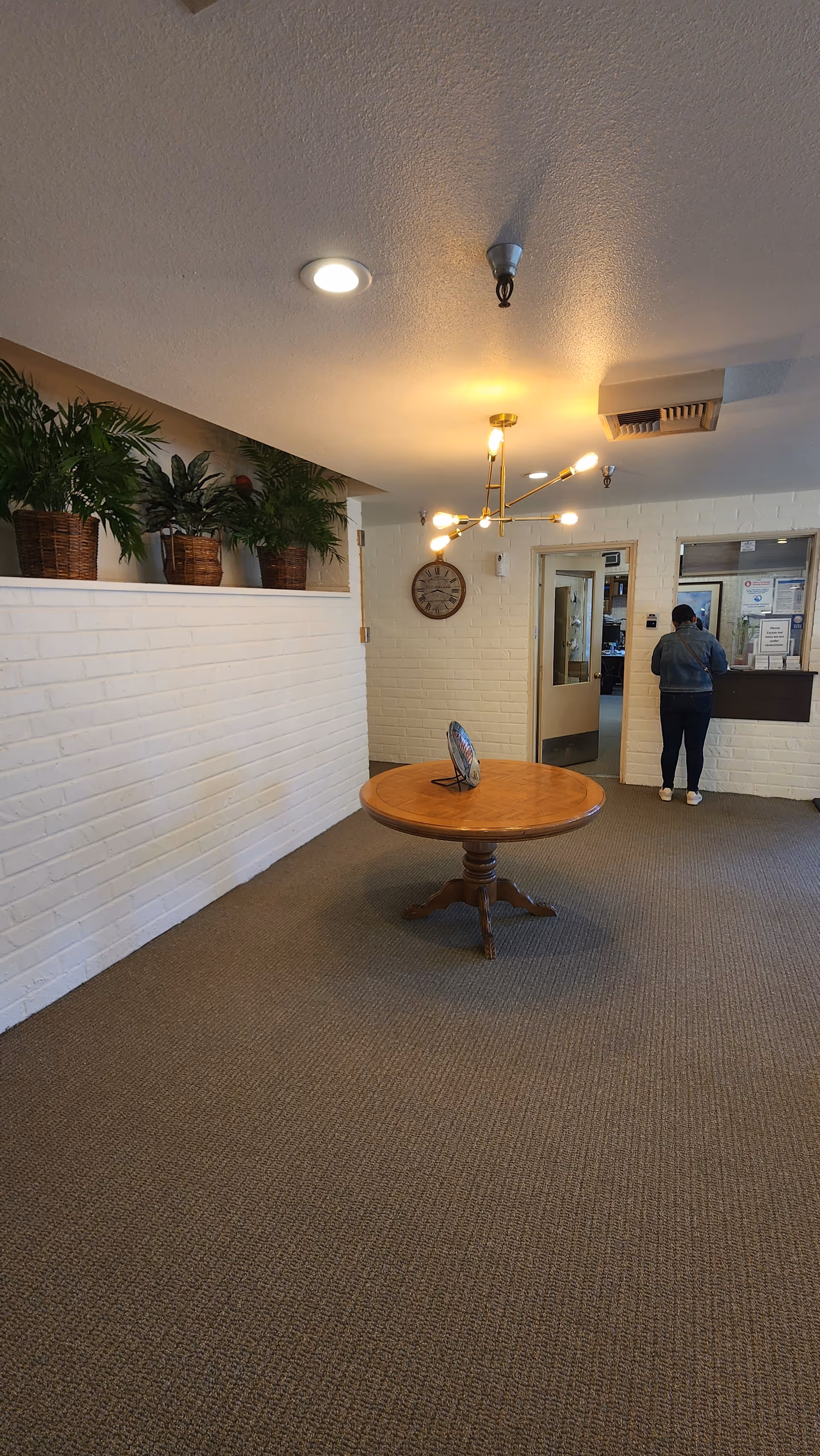 Interior view of a nursing home lobby or reception area with a round wooden table in the center, a modern chandelier overhead, potted plants on a ledge along a white brick wall, a clock on the wall, and a person standing at a reception window.