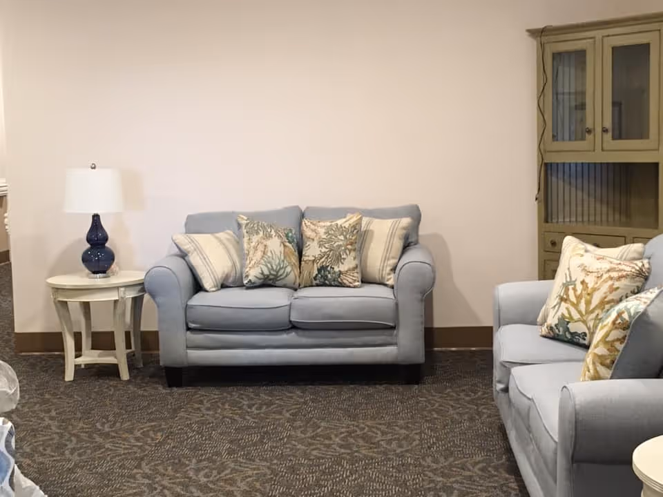 A cozy living room area with two light blue upholstered loveseats adorned with decorative pillows featuring coral and striped patterns. There is a small round white side table with a blue lamp on it to the left of one loveseat, and a tall cabinet with glass doors in the background on the right. The room has a patterned carpet and plain light-colored walls.