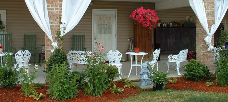 Outdoor patio area with white metal chairs and tables, green rocking chairs, potted plants, hanging pink flowers, and garden beds with various plants and mulch in front of a building with a white door and brick pillars.