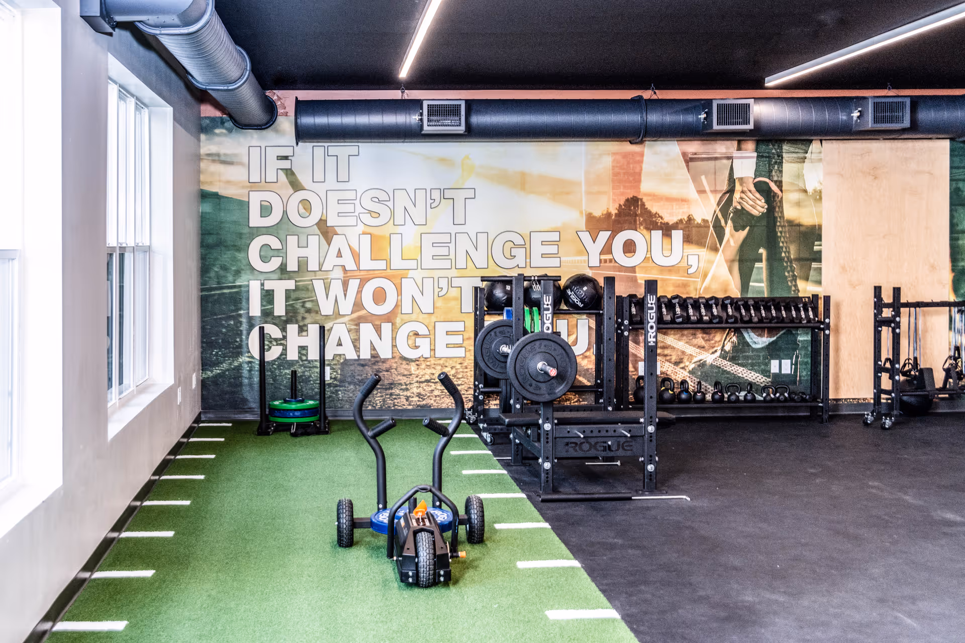 Interior view of a fitness room with green turf flooring and black rubber flooring. The room contains weightlifting equipment including barbells, kettlebells, and dumbbells. A motivational wall mural reads, 'IF IT DOESN'T CHALLENGE YOU, IT WON'T CHANGE YOU.'