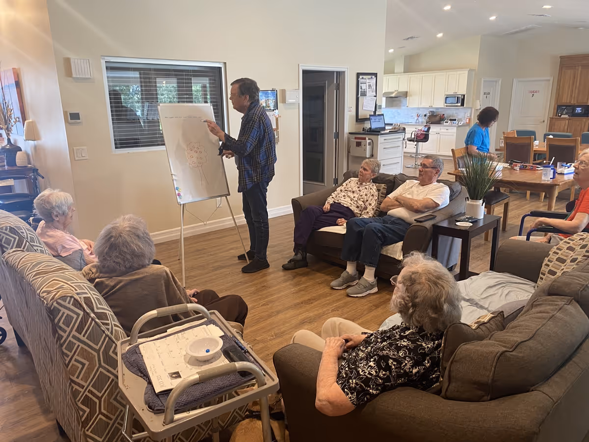 A group of elderly people seated on sofas and chairs in a living room area attentively watching a man standing and drawing on a whiteboard. The room has wooden flooring, a kitchen area in the background, and a woman working at a table.