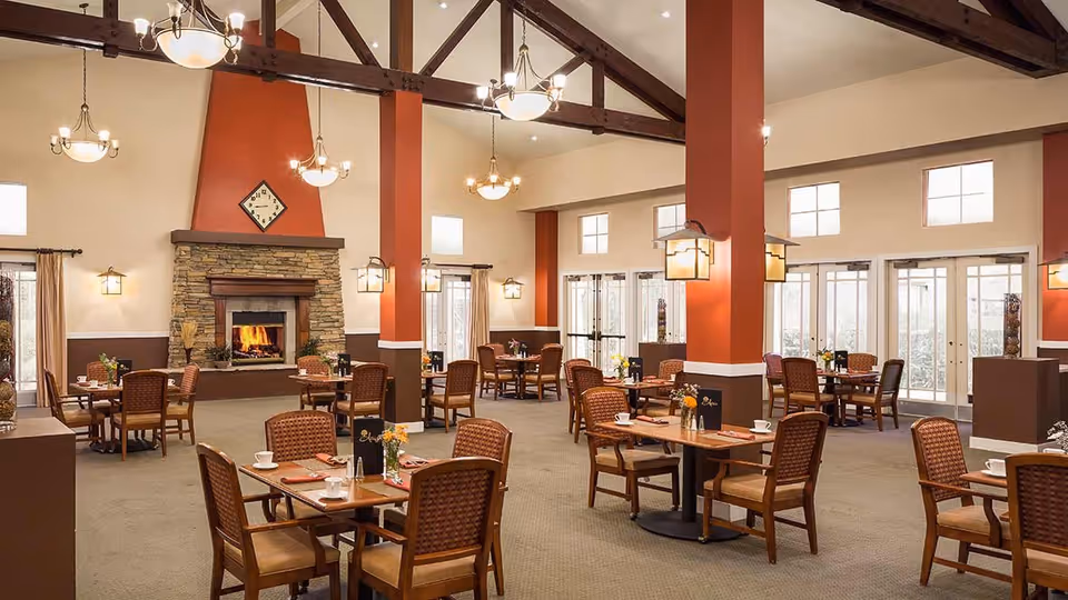 Spacious dining room with multiple wooden tables and chairs arranged neatly. The room features a stone fireplace with a clock above it, warm lighting from chandeliers and wall sconces, and large windows allowing natural light to enter.