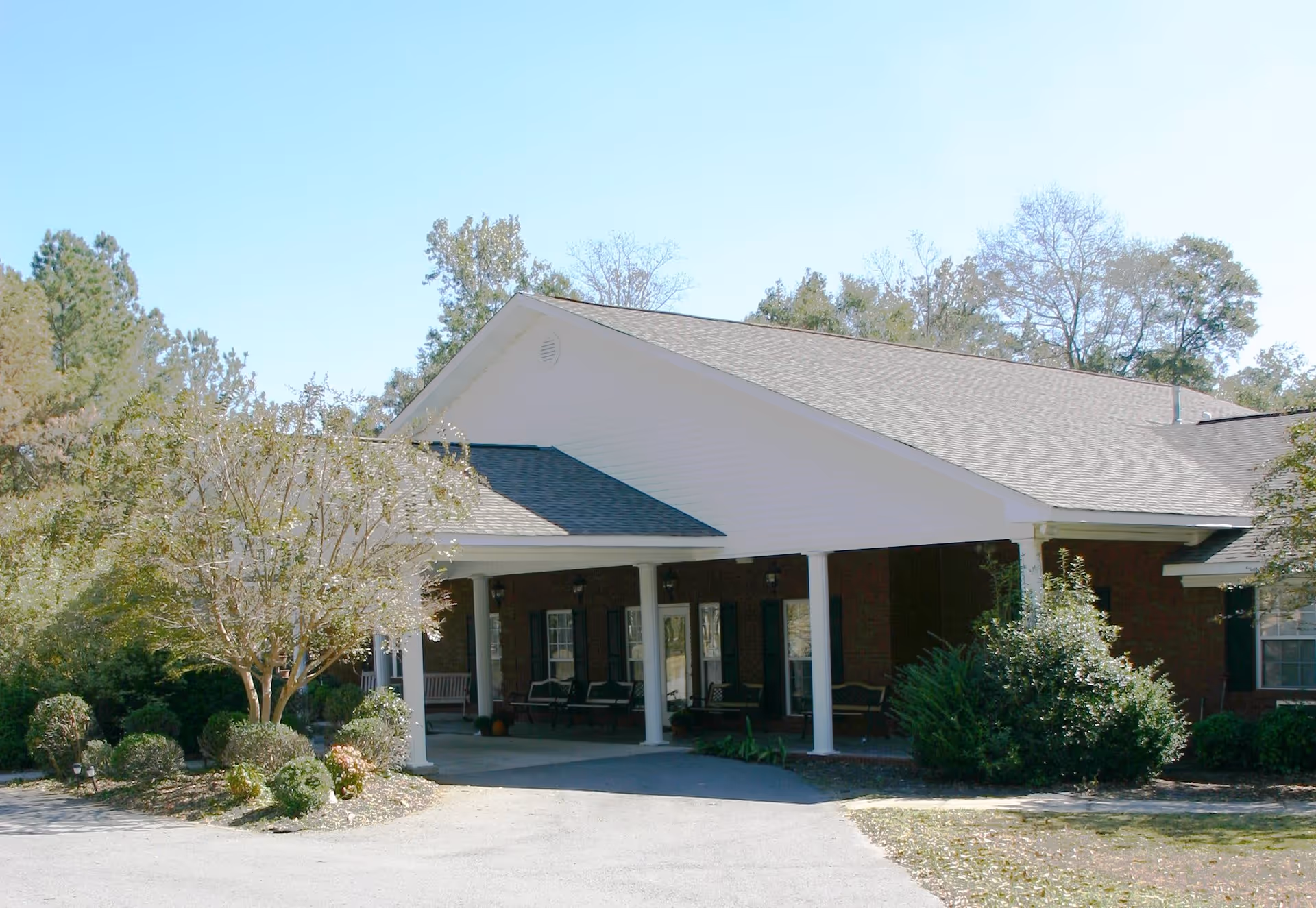 Exterior view of Morning Glory Meadows Assisted facility showing a single-story building with a covered entrance supported by white columns, surrounded by trees and shrubs under a clear blue sky.