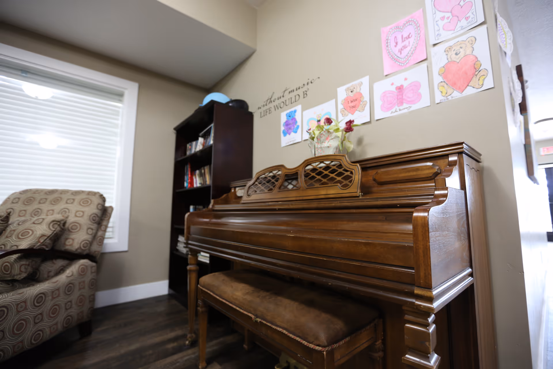 A cozy room featuring a wooden upright piano with a cushioned bench. Above the piano, children's drawings and Valentine-themed artwork are displayed on the wall. To the left, there is a patterned armchair and a dark wooden bookshelf filled with books. A window with white blinds is also visible.