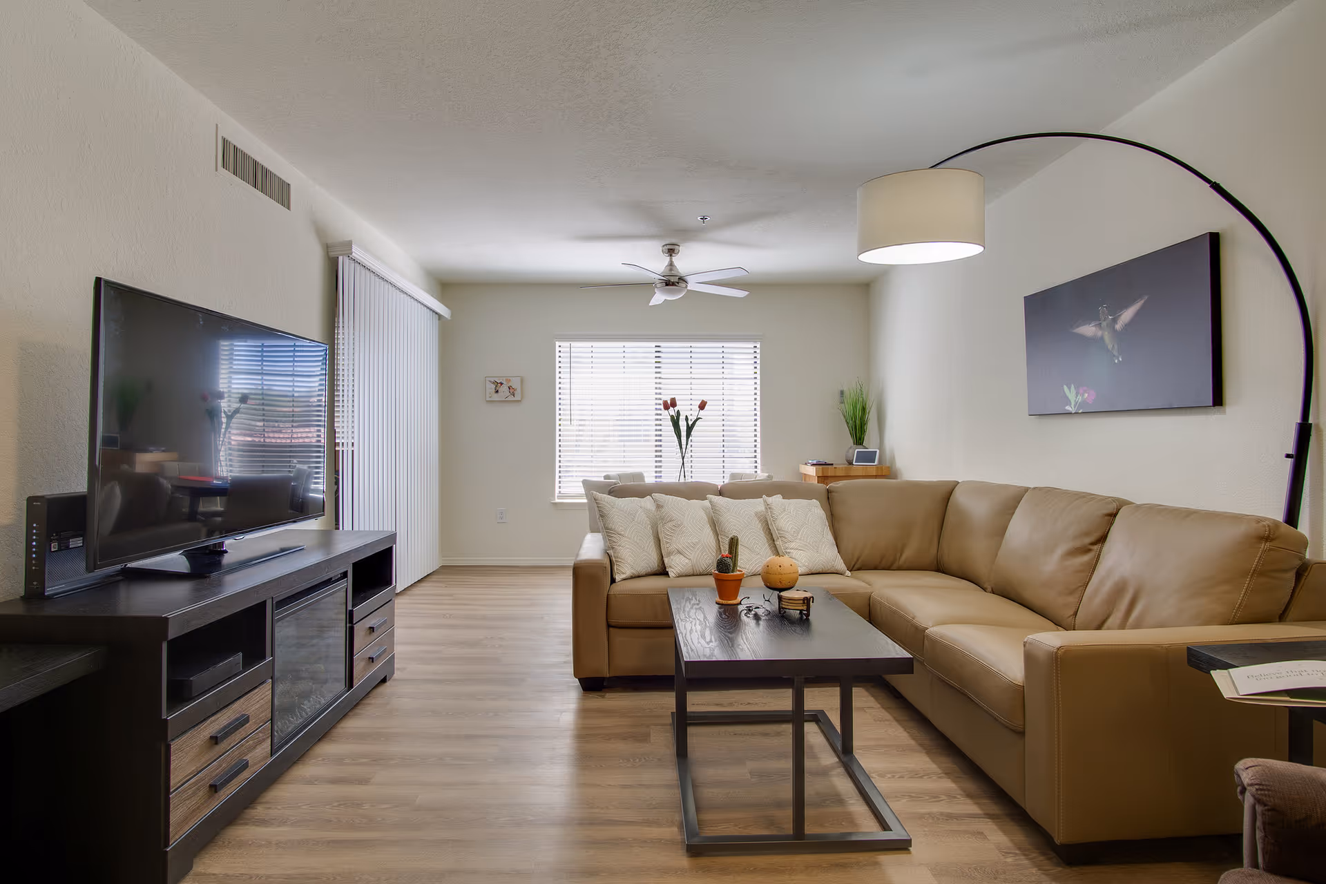 A modern living room with a tan leather sectional sofa adorned with white patterned pillows, a dark wooden coffee table with small decorative items, a large flat-screen TV on a dark wooden TV stand, a tall arc floor lamp, and a window with blinds letting in natural light. The room has light-colored walls and wood flooring.
