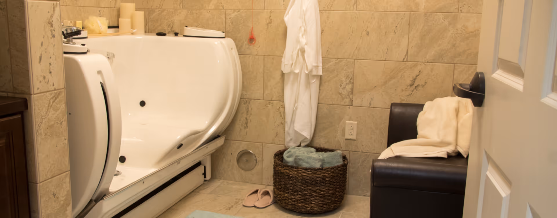 A bathroom with beige tiled walls and floor featuring a white walk-in bathtub with grab bars. There is a dark brown cushioned bench with a folded towel on it, a woven basket with rolled towels, a white bathrobe hanging on the wall, and a pair of pink slippers on the floor.