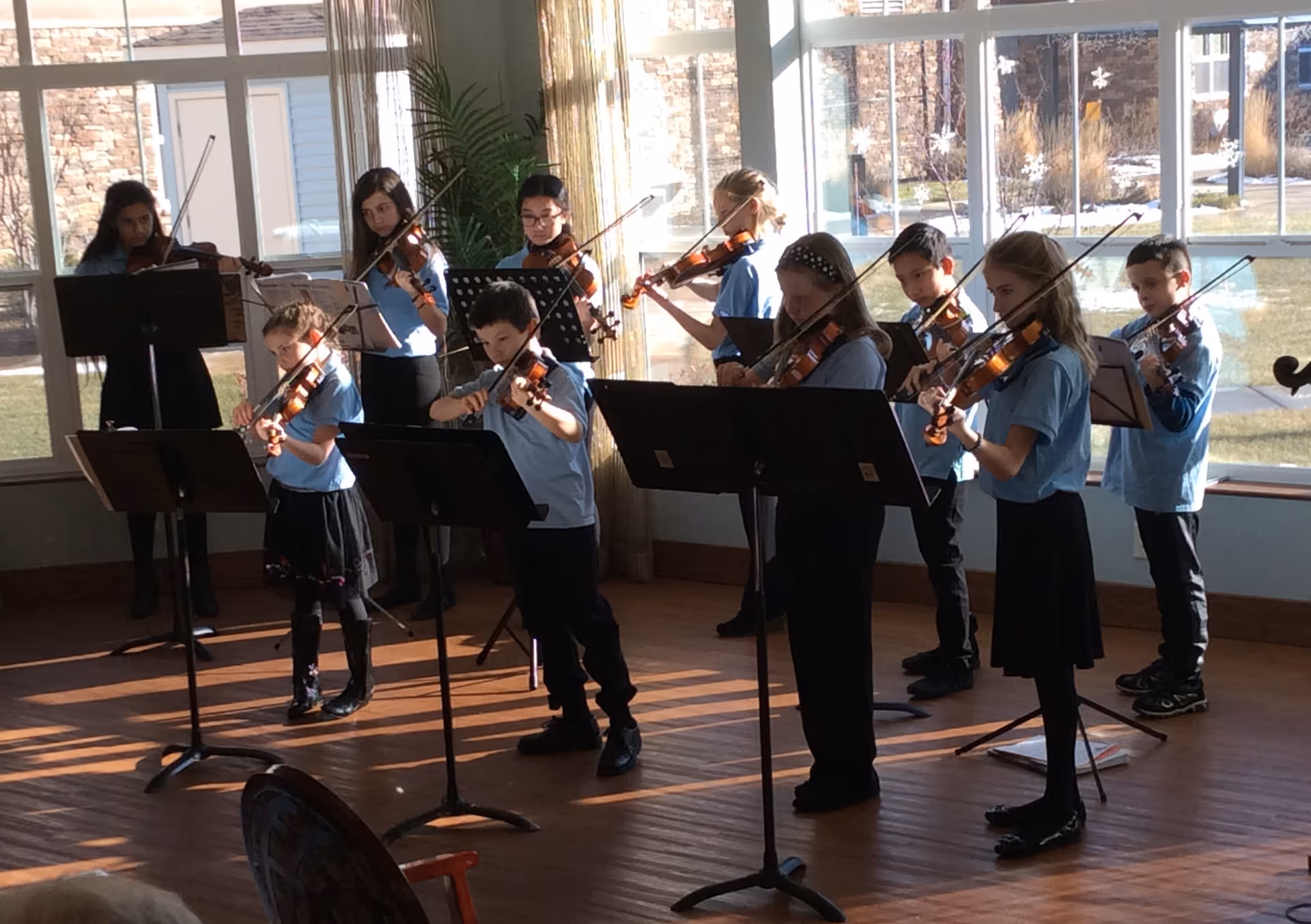 A group of children playing violins indoors in a sunlit room with large windows. They are standing in two rows, reading music from stands in front of them. The children are wearing light blue shirts and dark bottoms.