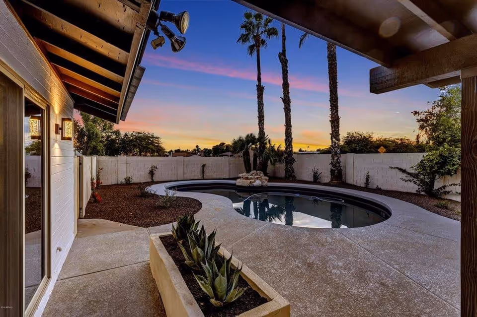 Outdoor view of a backyard at sunset featuring a kidney-shaped swimming pool surrounded by a concrete patio. There are tall palm trees and various plants along a white brick wall fence. The area is partially covered by a wooden roof with mounted lights.