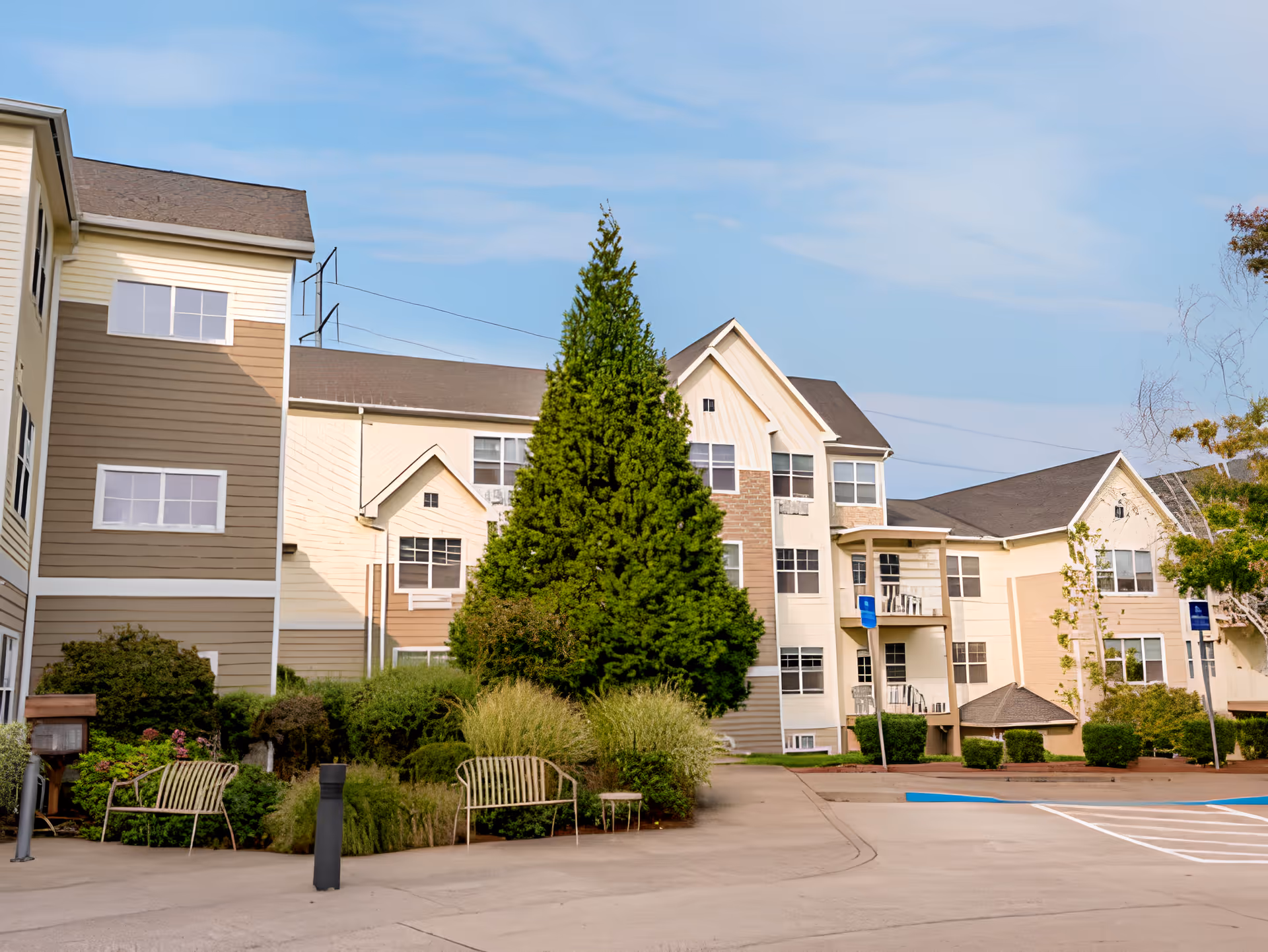 Exterior view of a multi-story senior living facility building with beige and light brown siding, multiple windows, and a pitched roof. The foreground features a paved parking area with marked handicap parking spaces, landscaped greenery including bushes and a tall evergreen tree, and two metal benches.
