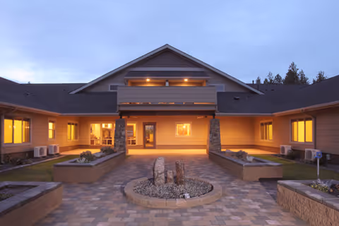 Exterior view of The Lodge at Riverside Harbor building at dusk with warm lights glowing from windows, a paved courtyard with planters, and a central circular stone feature.