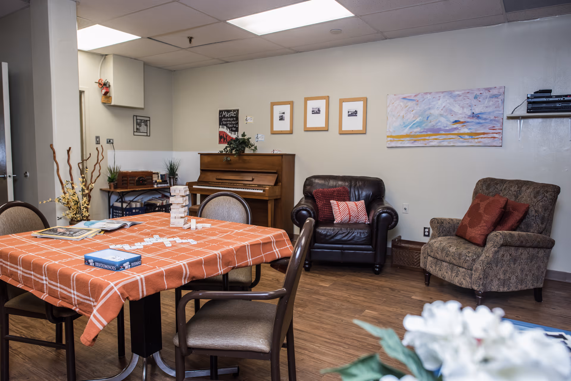 A cozy common room with a checkered table and chairs, a piano against the wall, and two upholstered armchairs.
