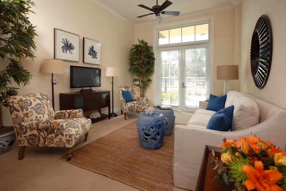 A cozy living room with a beige sofa adorned with blue pillows, two patterned armchairs, a dark wooden TV stand with a flat-screen TV, two floor lamps, a large window with blinds and a glass door leading outside, two blue ceramic stools as a coffee table, a large round decorative mirror on the wall, and a vase of orange flowers on a side table.