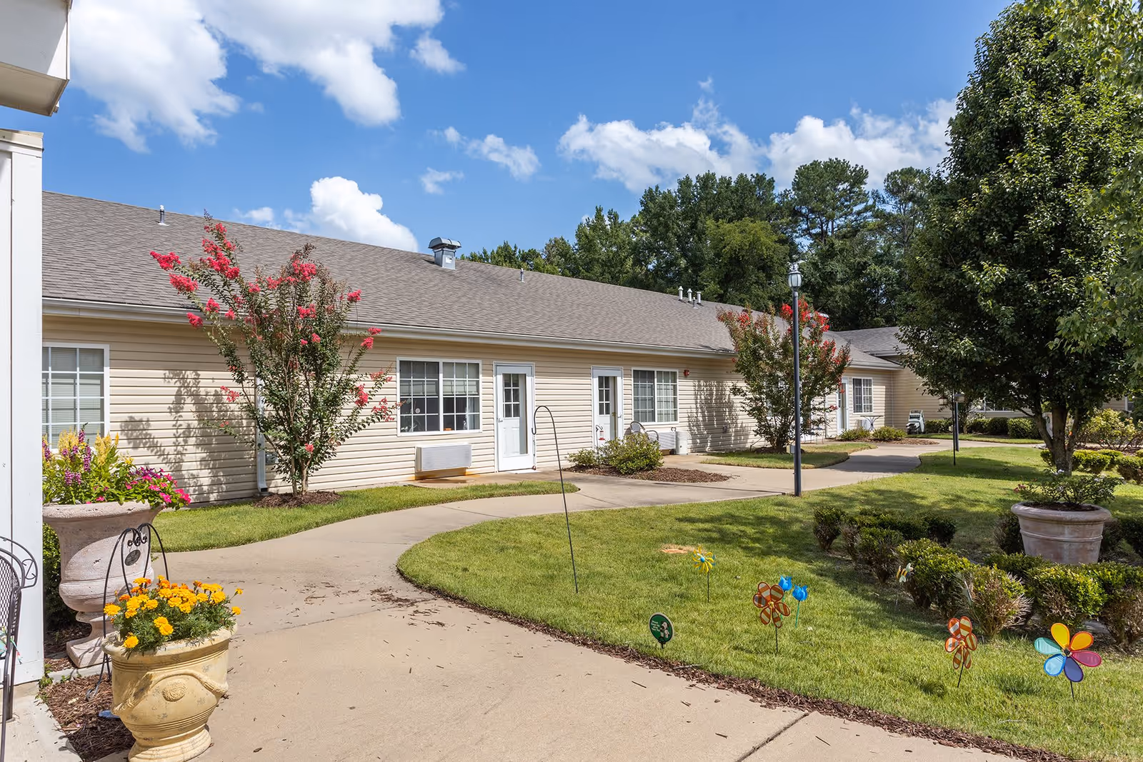 Outdoor view of a single-story assisted living facility building with beige siding and white doors and windows. The area features a curved concrete walkway, green grass, colorful flower pots, small bushes, and decorative pinwheels on the lawn. Trees and a bright blue sky with scattered clouds are visible in the background.
