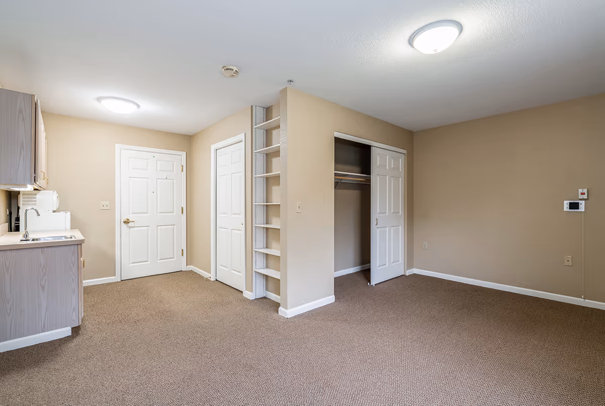 An empty interior room in a senior living facility featuring beige walls and carpeted floor. The room includes a small kitchenette with a sink, microwave, and cabinets on the left side. There are two white doors in the center, one likely an entrance door and the other a closet door. To the right, there is an open closet with sliding doors and a small built-in shelving unit between the doors. The ceiling has two round light fixtures.