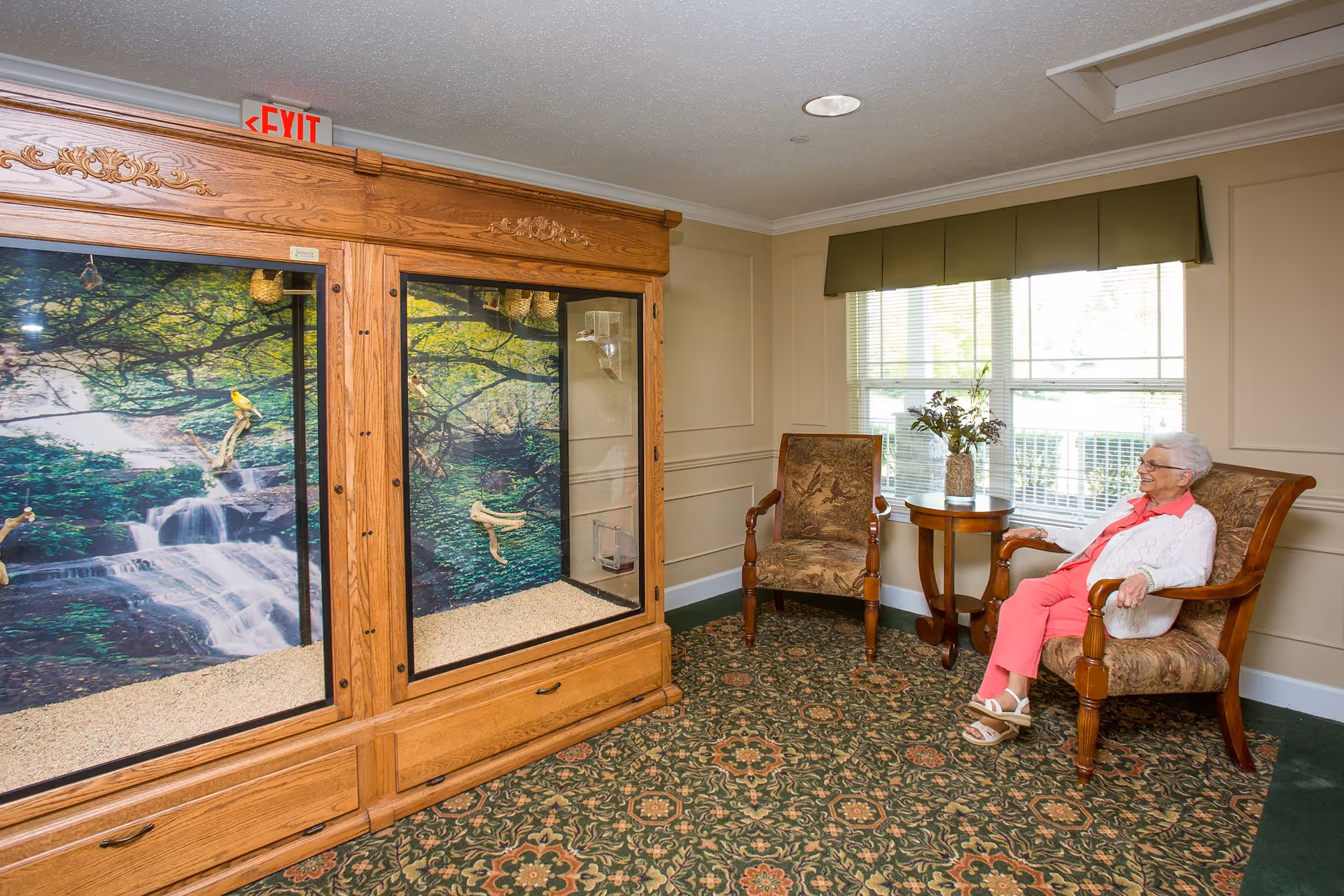 An elderly woman sitting on a cushioned wooden chair in a cozy room with patterned carpet and beige walls. Next to her is a small round wooden table with a vase of flowers. Across from her is a large wooden bird enclosure with a nature-themed backdrop featuring a waterfall and trees. Another cushioned wooden chair is positioned near the window with blinds and a green valance.