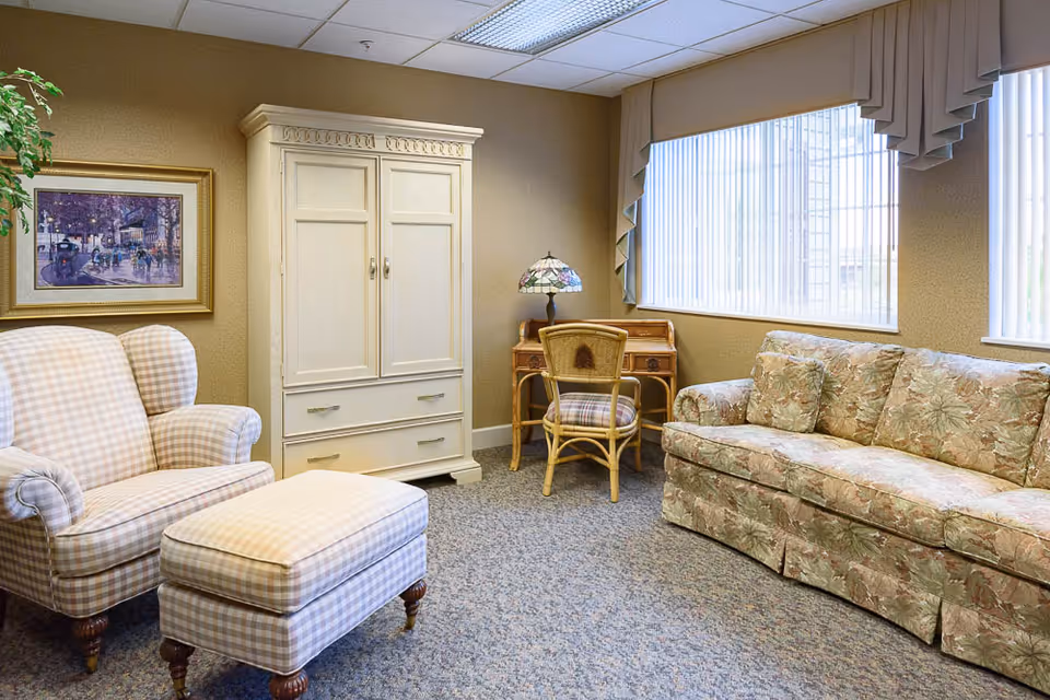 A cozy living room with a floral patterned sofa, a checkered armchair with matching ottoman, a white wooden cabinet, a small wooden desk with a wicker chair, and a Tiffany-style lamp. The room has beige walls, a large window with vertical blinds, and a framed painting on the wall.