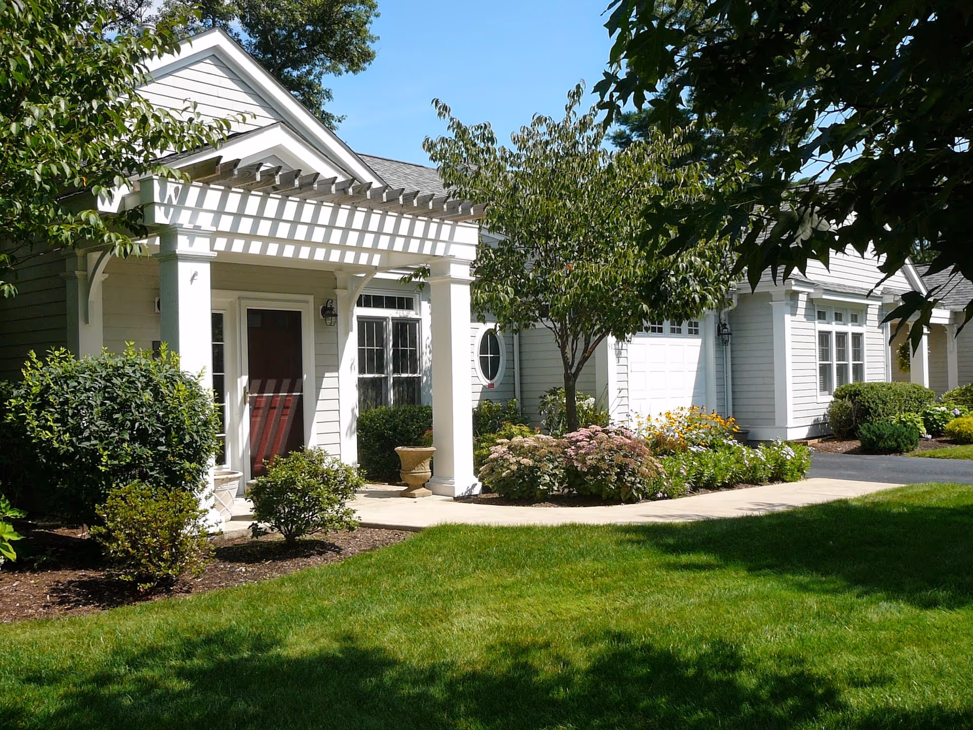 Exterior view of a white residential building with a small porch supported by columns and a pergola. The building is surrounded by well-maintained green grass, bushes, flowering plants, and trees under a clear blue sky.
