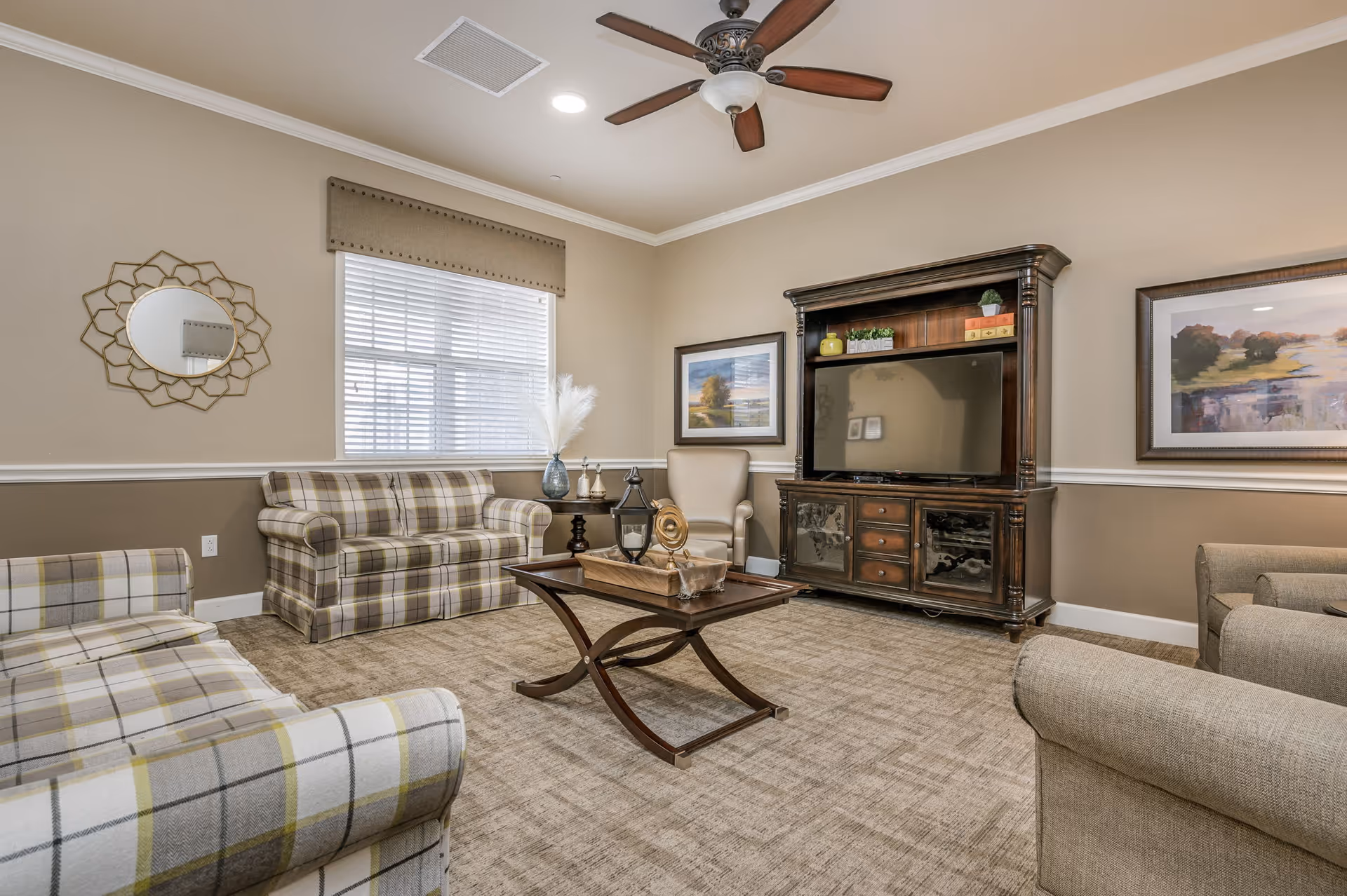 A cozy living room with beige walls and carpet. The room features a plaid sofa and loveseat, a beige armchair, a wooden coffee table with decorative items, and a large dark wood entertainment center with a flat-screen TV. There are two framed landscape paintings on the walls and a window with white blinds and a beige valance. A ceiling fan with lights is mounted on the ceiling.