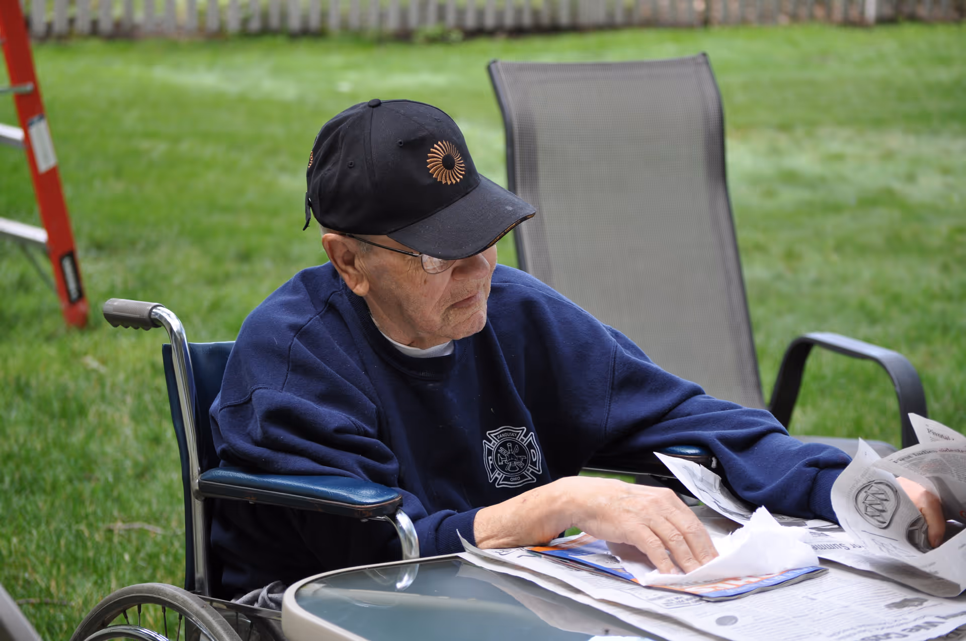 An elderly man in a wheelchair wearing a black cap and navy blue sweatshirt is sitting outdoors at a glass table, reading and sorting through newspapers and papers. Behind him is a lawn chair and green grass.