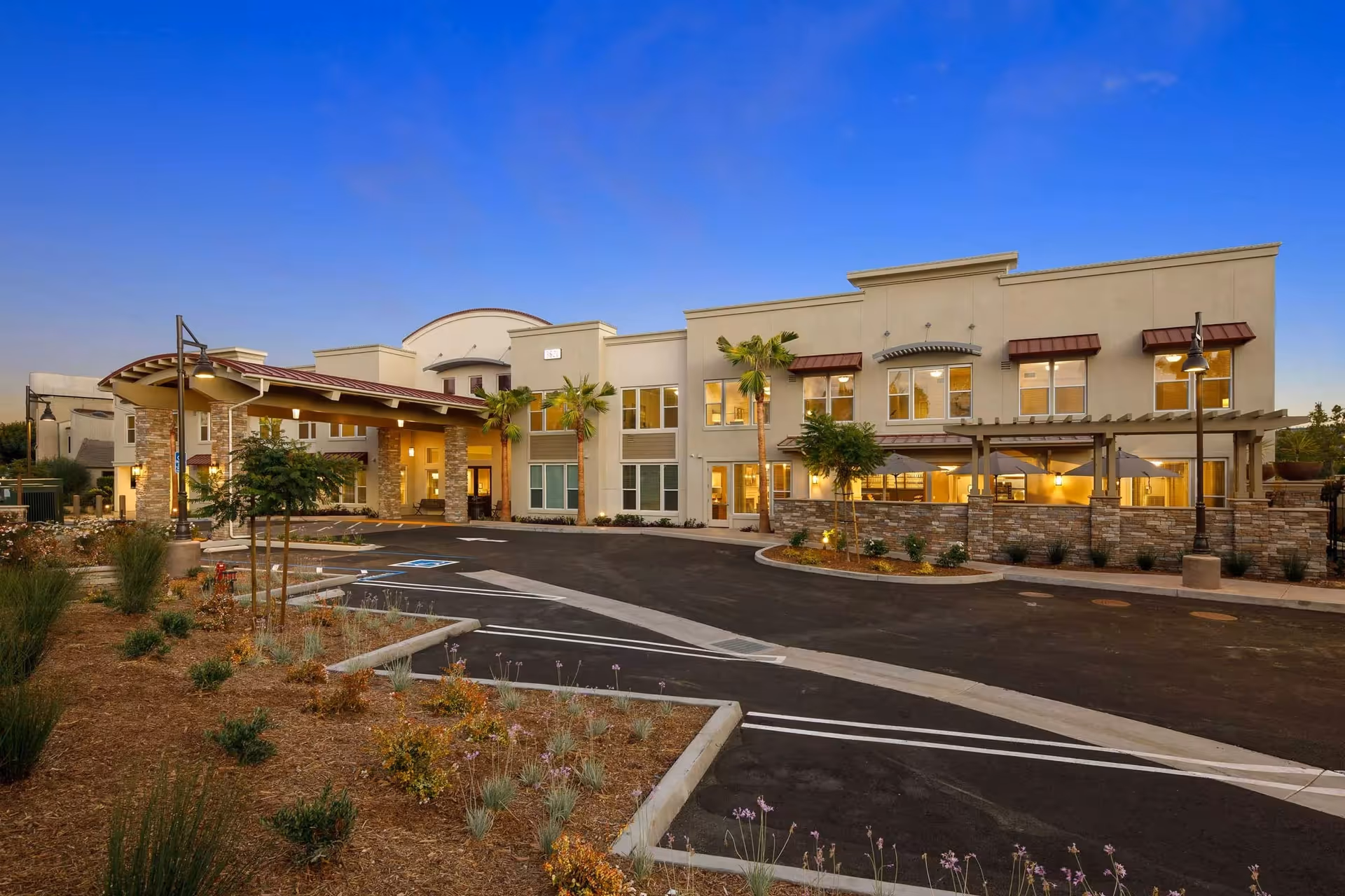 Exterior front entrance of a two-story senior living facility at dusk with a covered porte-cochère, landscaped beds, and a parking/drop-off area.