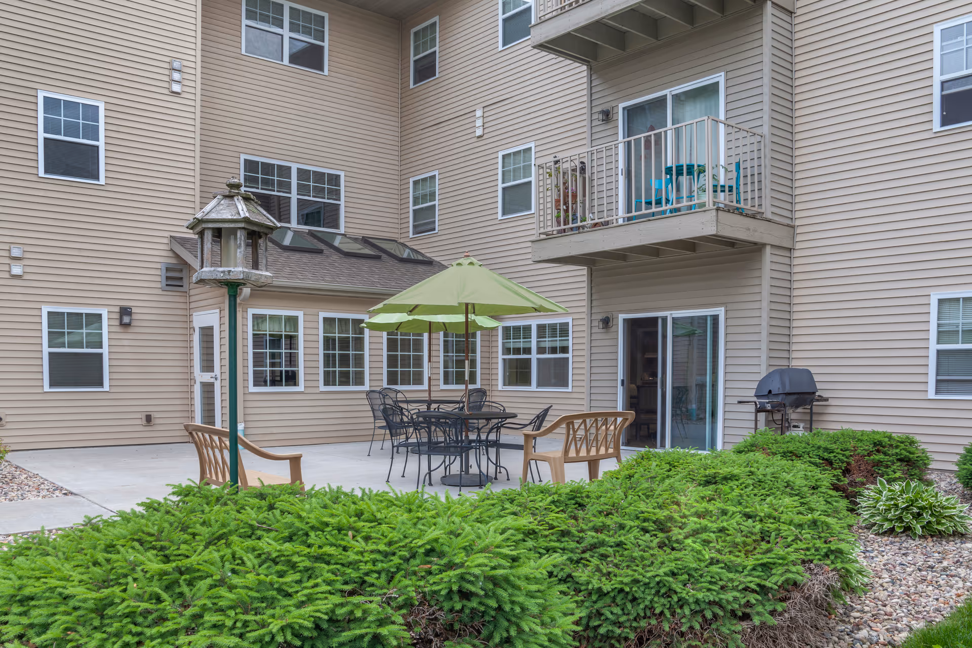 Outdoor courtyard patio at a multi-story residential building with a table and green umbrella, chairs, grill, and surrounding shrubs.