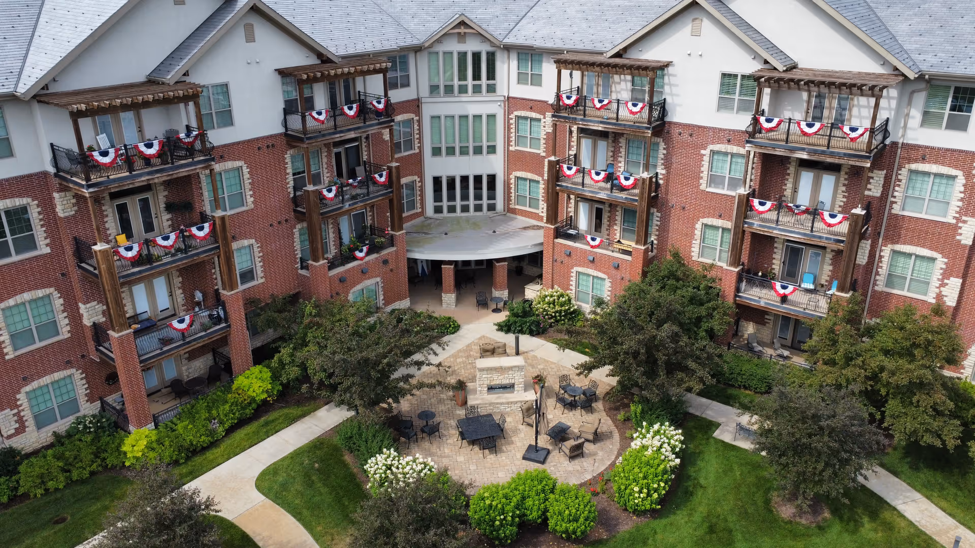 Aerial view of a brick senior living building with balconies decorated with red, white, and blue bunting overlooking a landscaped courtyard with patio seating.