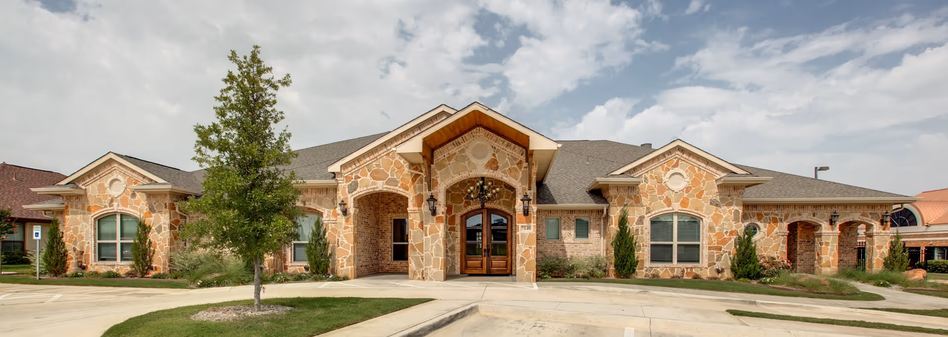 One-story stone-faced memory care building with an arched main entrance, windows, landscaping, and a parking area under a partly cloudy sky.