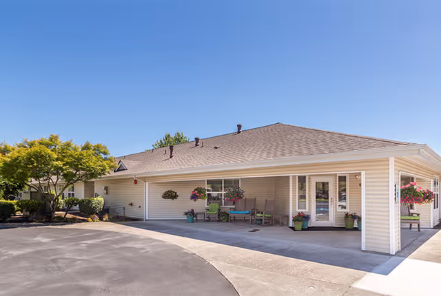 Exterior view of a single-story building with beige siding and a gray shingled roof under a clear blue sky. The building has a covered patio area with hanging flower baskets and outdoor seating. There are trees and shrubs around the building and a paved driveway in front.