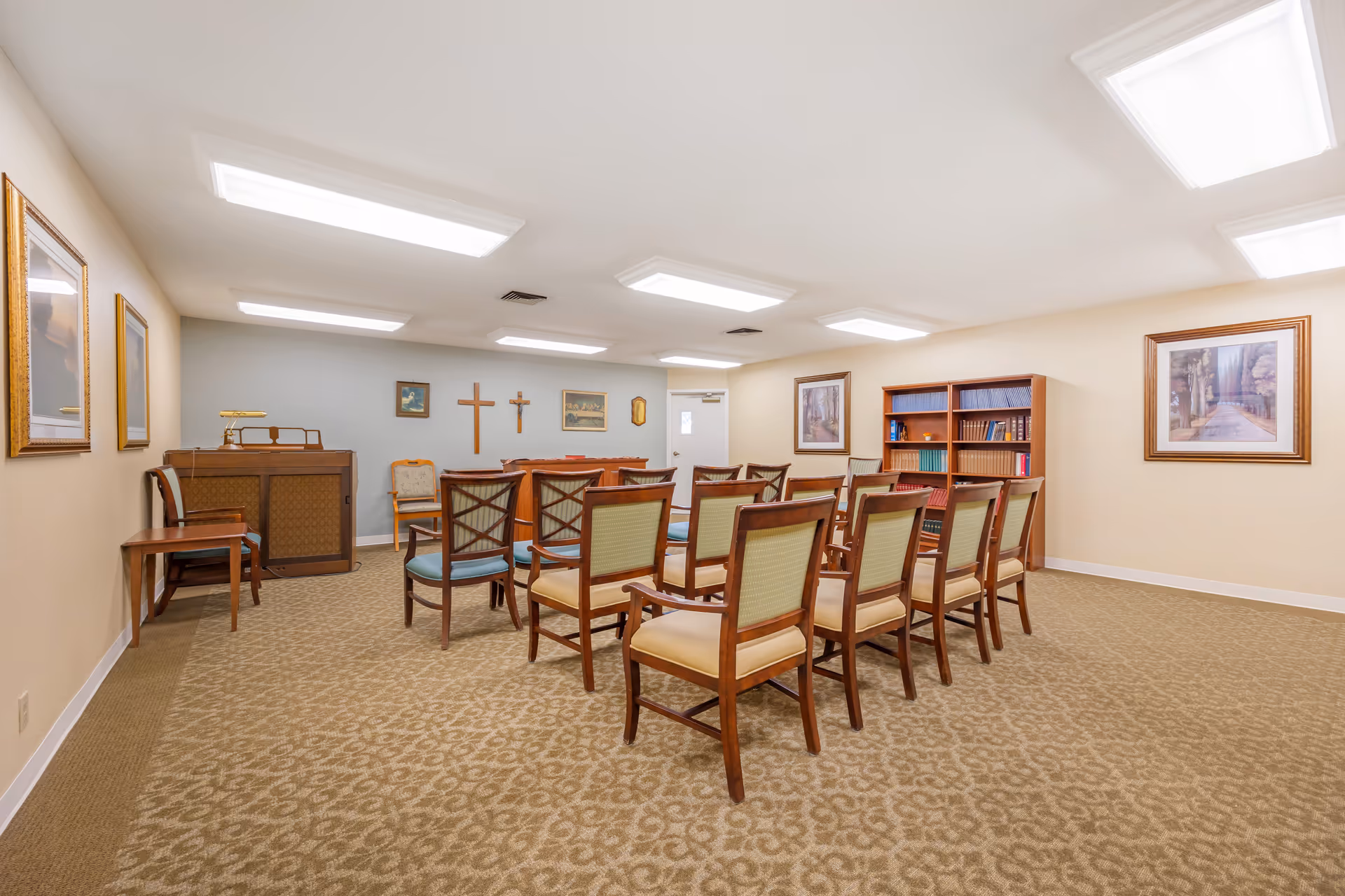 A small chapel or meeting room with rows of wooden chairs facing a wooden podium. The room has beige walls, carpeted floor, framed pictures on the walls, and two crosses mounted on the far wall. There is a bookshelf filled with books and a wooden organ or piano on the left side of the room.