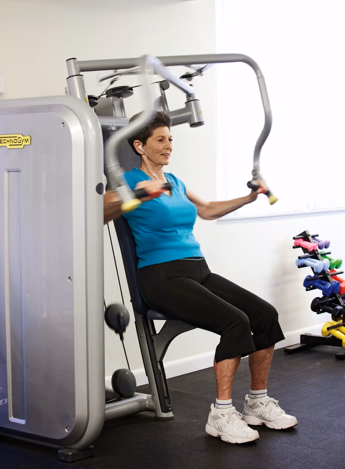 An elderly woman wearing a blue sleeveless top and black pants is exercising on a chest press machine in a fitness room. She is seated and pushing the machine's handles forward. In the background, there is a rack with colorful dumbbells and a window letting in natural light.