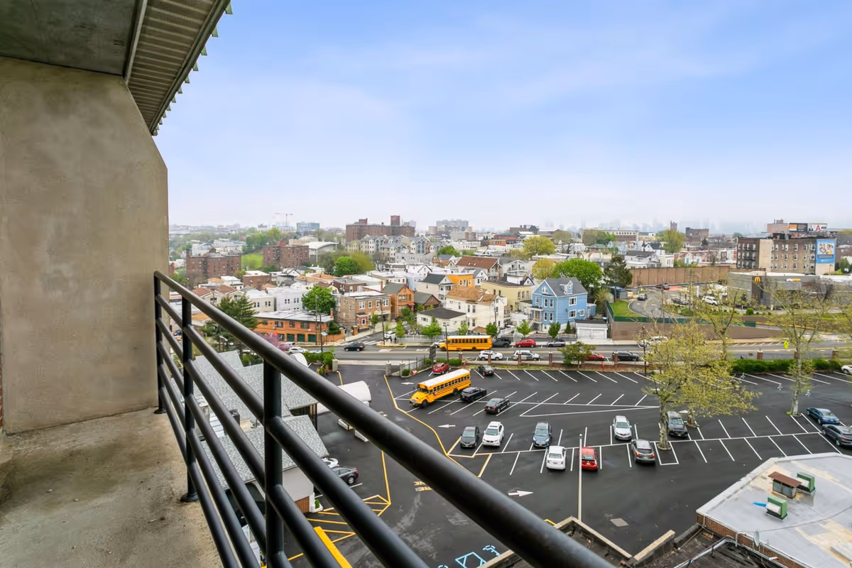 View from a balcony overlooking a parking lot with several cars and two yellow school buses, with a neighborhood of houses and buildings in the background under a cloudy sky.