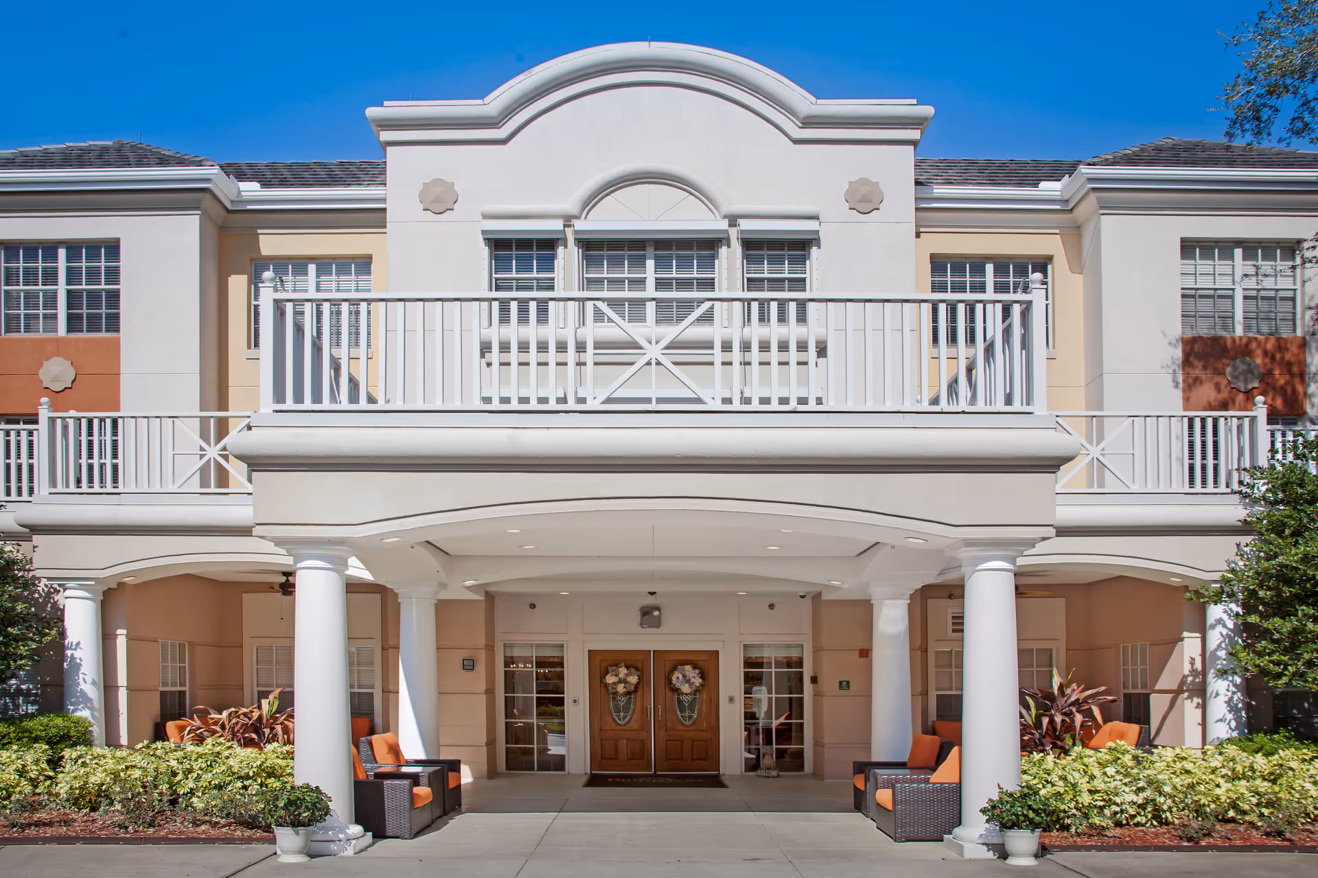 Front entrance of a two-story senior living building with white columns, an upper balcony, and outdoor seating.