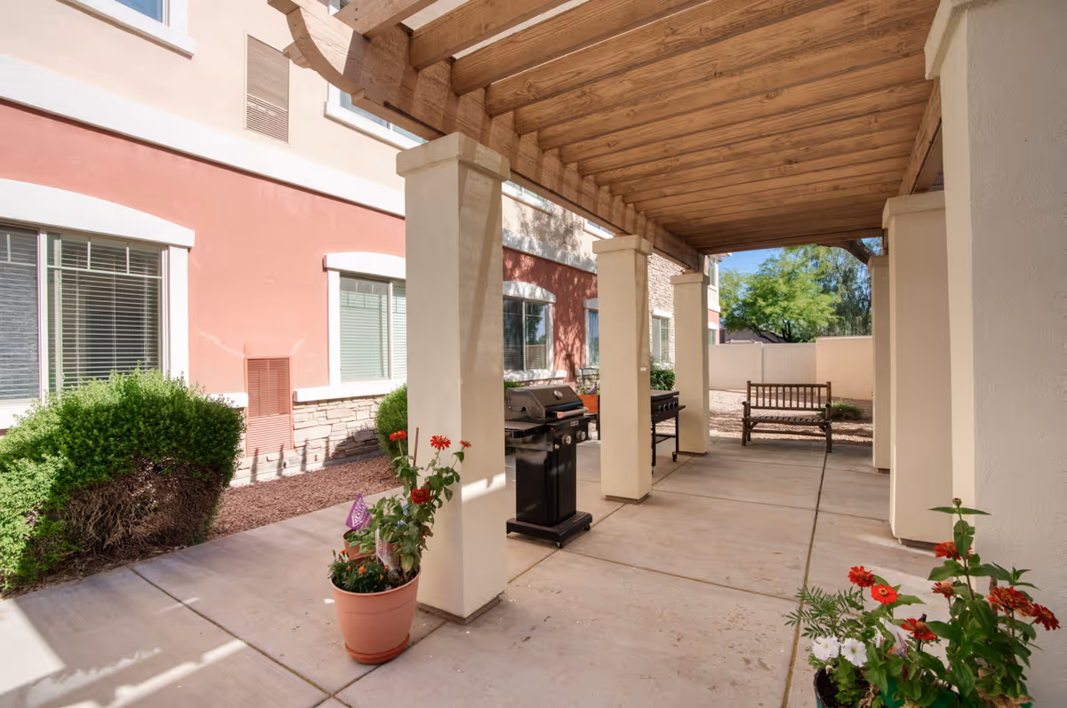 Covered outdoor patio with a wooden pergola, columns, grills, benches, and potted plants alongside a multi-story building.