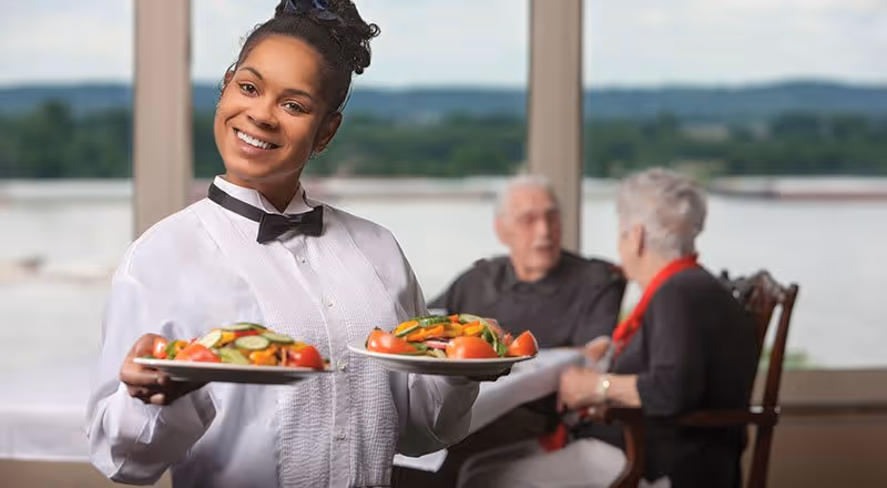 A smiling waitress dressed in a white shirt and black bow tie holding two plates of fresh salad. In the background, an elderly man and woman are seated at a table near large windows with a scenic view outside.