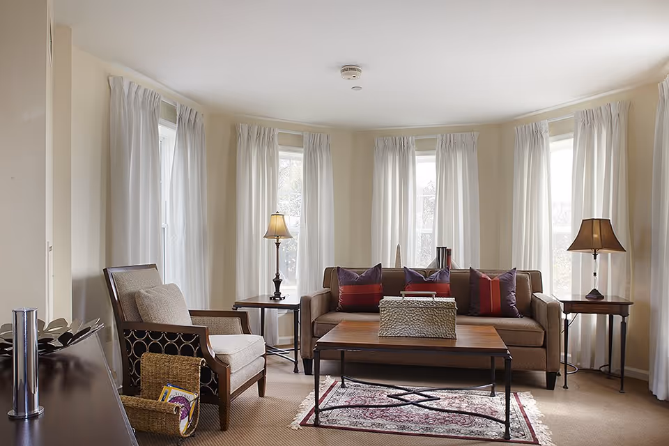 A cozy living room with a brown sofa adorned with red and purple pillows, a wooden coffee table with a decorative basket, two side tables each with a lamp, a cushioned armchair, and large windows with white curtains letting in natural light.