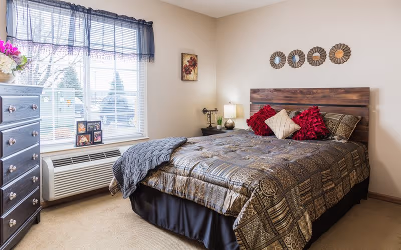 A furnished bedroom with a made bed featuring patterned bedding and red accent pillows, a wooden headboard and dresser, bedside lamp, and a window with blinds.