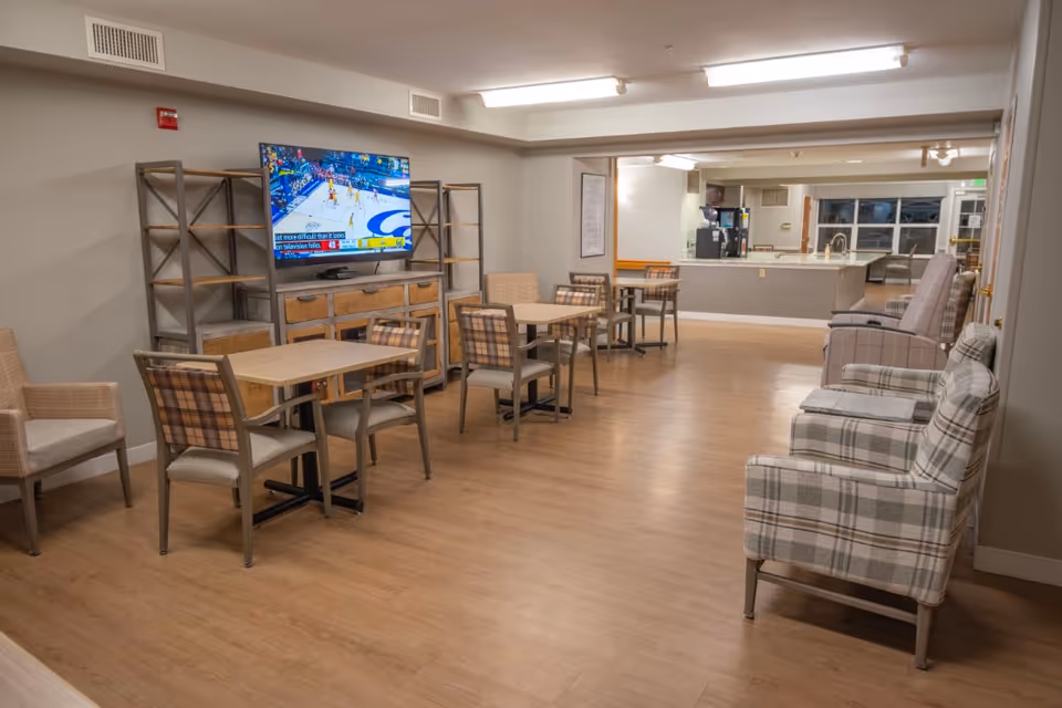 A common area in a senior living facility featuring several small tables with chairs, a television mounted on a wooden shelving unit showing a basketball game, and multiple upholstered armchairs arranged along the walls. The room has wood flooring and neutral-colored walls, with a kitchen or service area visible in the background.