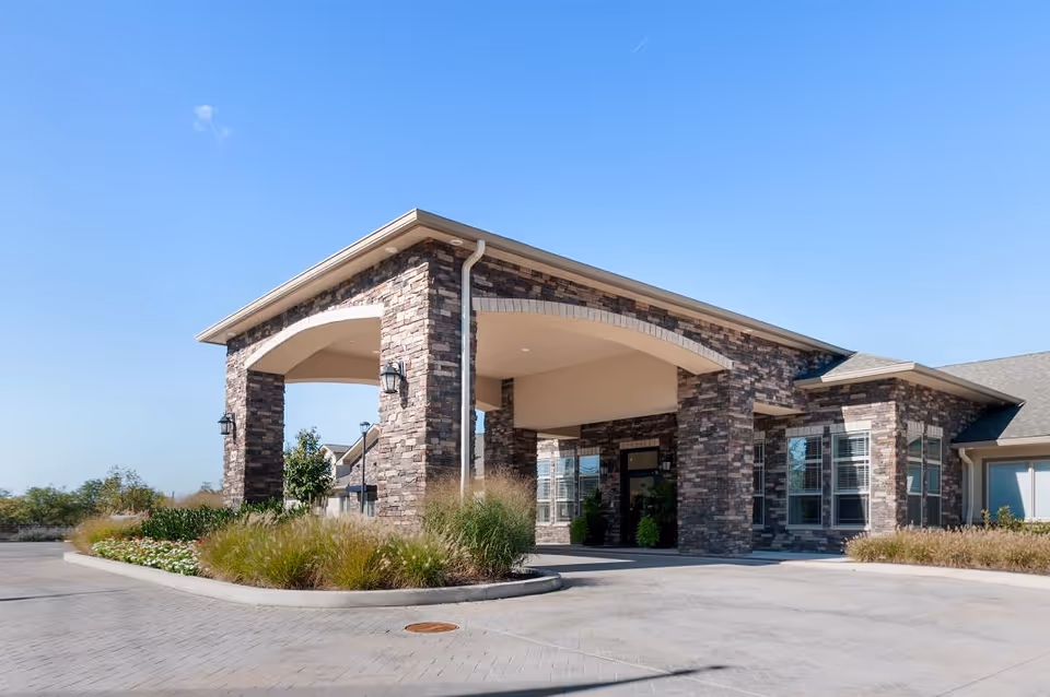 Exterior view of a senior living facility entrance with a covered drop-off area supported by stone pillars, surrounded by landscaped greenery and clear blue sky.