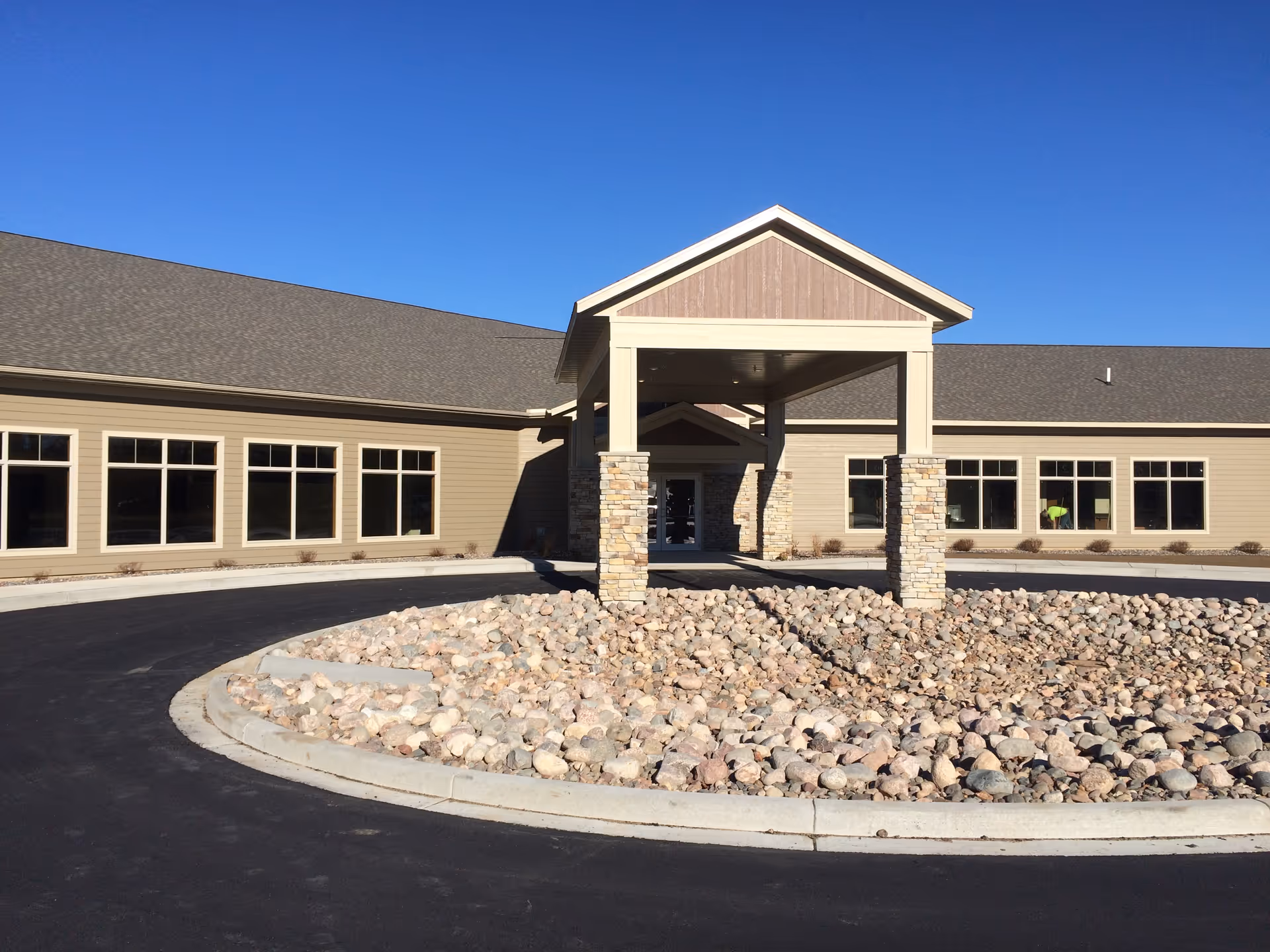 Front exterior view of Dove Healthcare - Bloomer building with a covered entrance supported by stone pillars, surrounded by a circular driveway and a landscaped area filled with rocks under a clear blue sky.