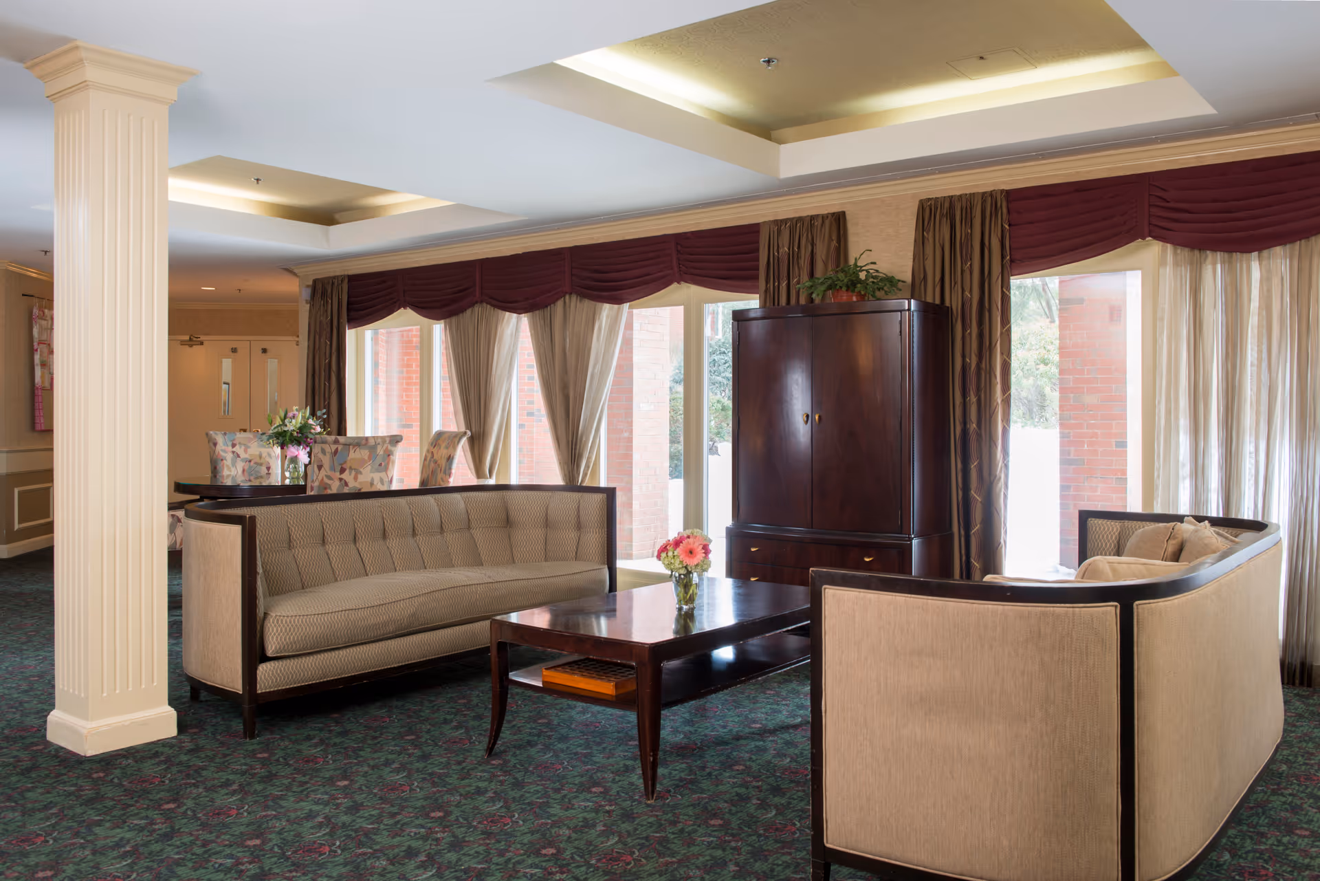 A cozy living room area with two beige curved sofas facing a dark wooden coffee table with a small vase of flowers. Behind the sofas is a dark wooden cabinet with a plant on top. Large windows with sheer curtains and burgundy valances allow natural light to fill the room. A dining table with floral upholstered chairs is visible in the background.