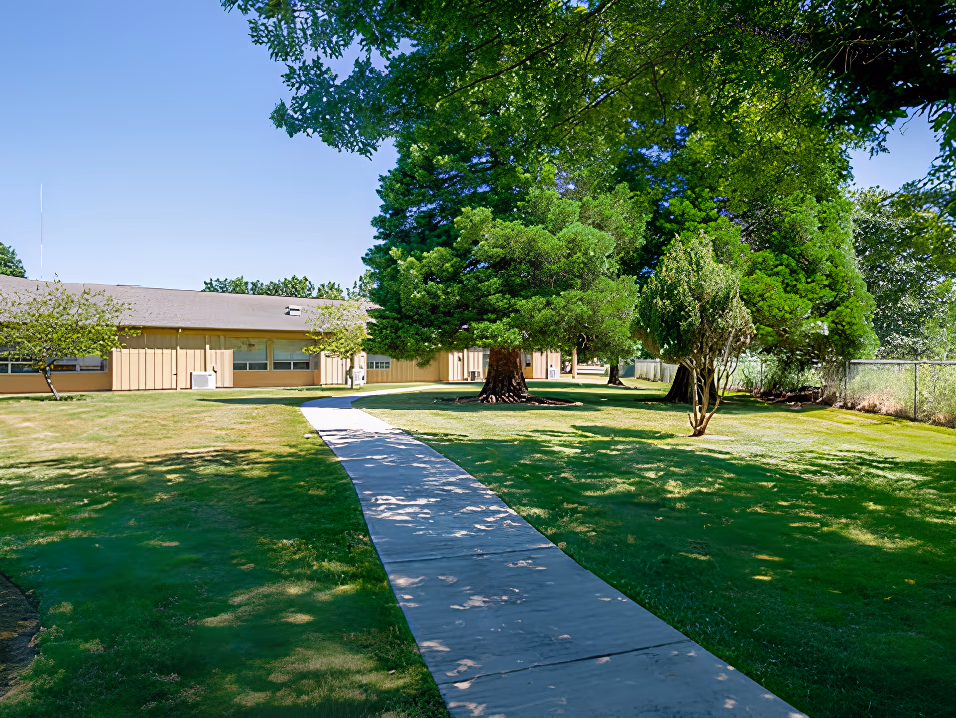 A paved walkway leading through a grassy outdoor area with several large green trees and a beige building in the background under a clear blue sky.