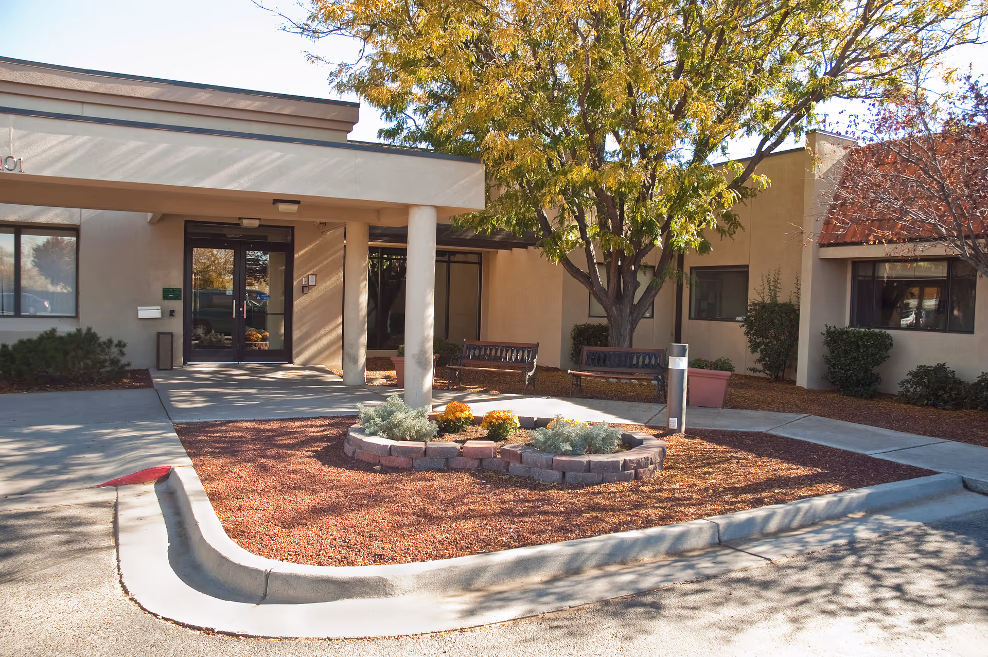 Entrance of a senior living facility with a covered walkway supported by columns, two benches, a small landscaped area with plants and flowers, and trees with autumn foliage.