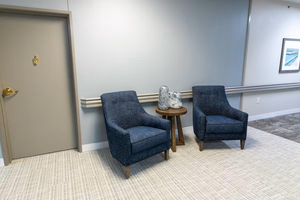 Two blue upholstered armchairs with wooden legs are placed on a light-colored carpeted floor. Between the chairs is a small round wooden side table holding two decorative marble vases. To the left is a closed beige door with a gold handle and a gold number 6. The walls are painted light gray with a chair rail molding, and a framed artwork is visible on the right wall.