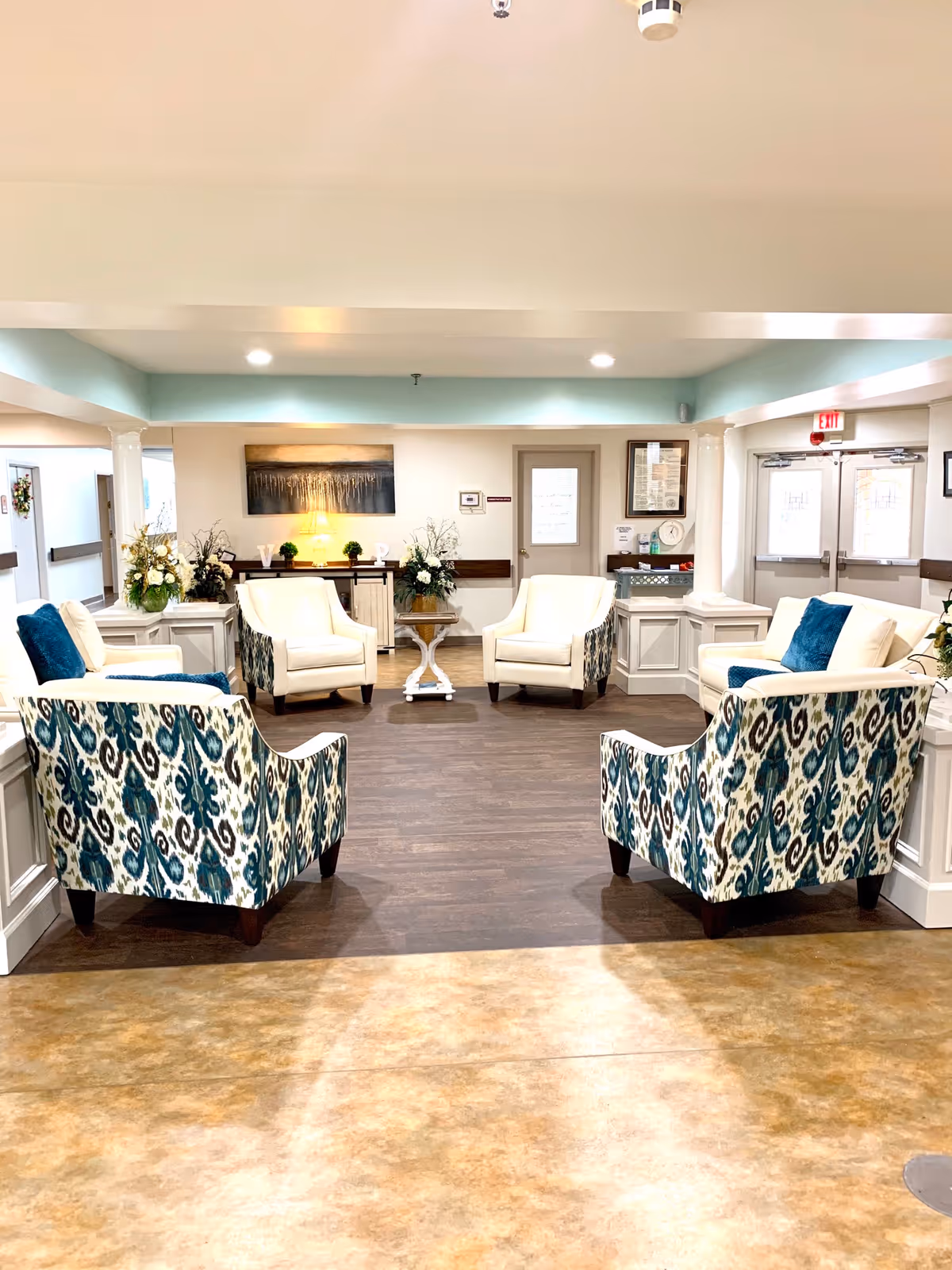 A bright and spacious common area with six armchairs arranged in a circle. Four chairs have a blue and white patterned upholstery, while two are solid white. The room has wood flooring in the seating area and a lighter tile floor in the foreground. There are decorative plants and flowers on side tables and a console table against the back wall, which also features a large piece of artwork. Double exit doors are visible on the right side of the room.