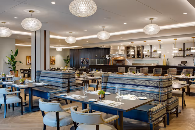 A spacious and modern dining room with multiple tables and chairs arranged neatly. The tables are set with white napkins, plates, and glassware. There are patterned upholstered bench seats and light blue cushioned chairs. The ceiling features round pendant lights, and the back wall has a counter area with bar stools and shelves with decorative items and lighting.