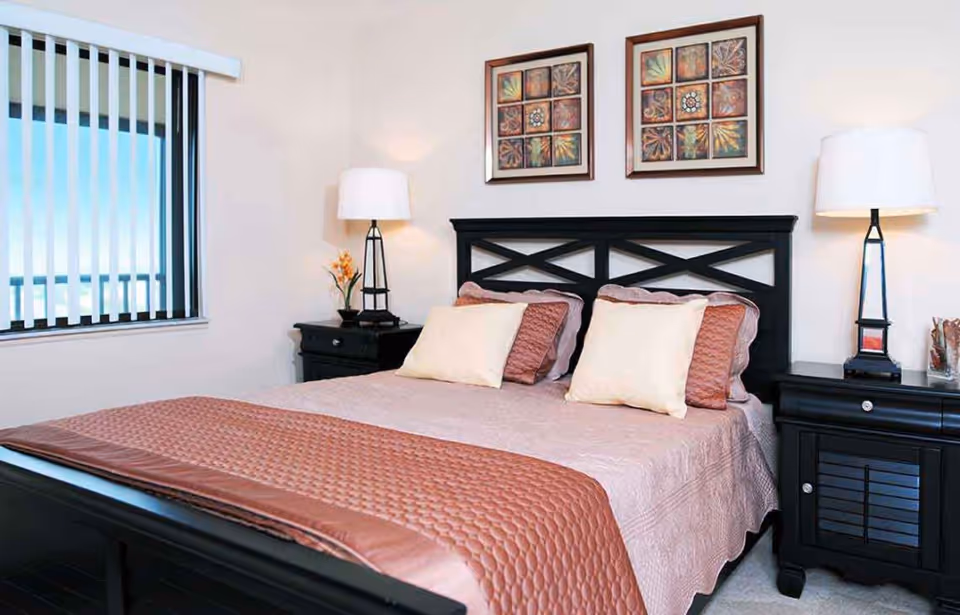 A neatly made bed with a black wooden headboard and footboard, covered with a light pink quilt and a textured brown throw blanket. There are four pillows, two cream-colored and two brown, arranged on the bed. On each side of the bed are black nightstands with drawers, each holding a modern lamp with a white shade. Above the bed are two framed pieces of artwork featuring abstract designs. A window with vertical blinds is visible on the left side of the room.